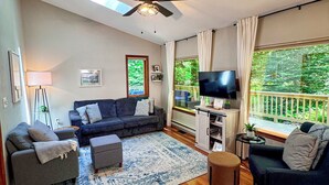 Ein Wohnzimmer mit Sofa, Hocker, Fernseher, Deckenventilator und großen Fenstern.
