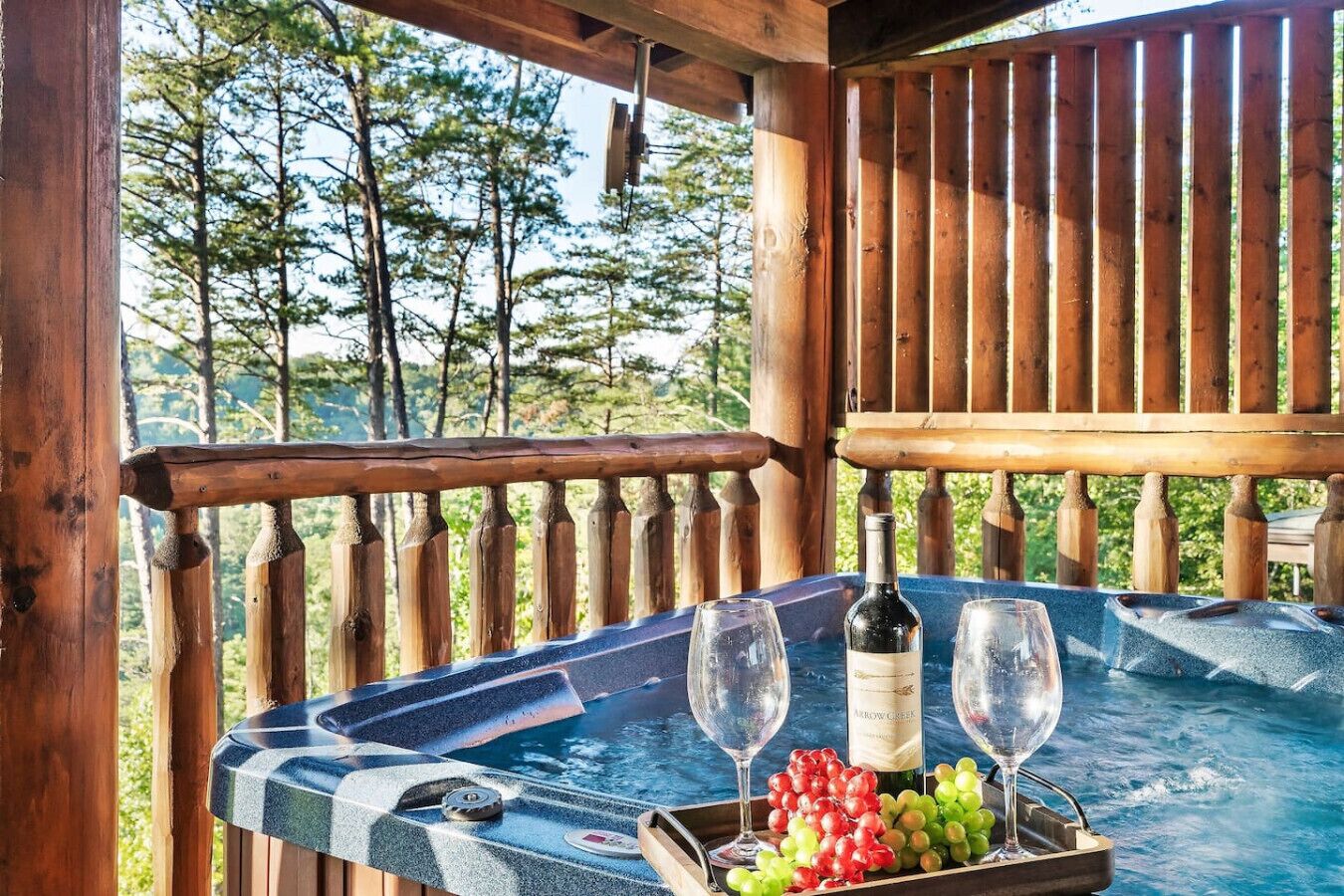 A wooden deck with a table set for a meal, featuring a bottle of wine and glasses, and a tray of grapes.