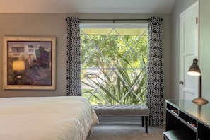 Quarto com uma cama, uma escrivaninha com abajur, uma janela com cortinas estampadas e um quadro na parede.