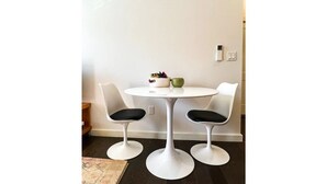 Una mesa pequeña y redonda con dos sillas blancas, una taza verde y una cesta de objetos sobre la mesa.
