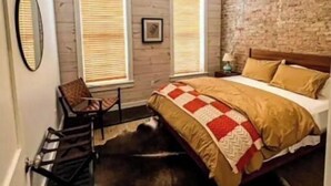 Un dormitorio con techo de madera, una cama con colcha, dos ventanas con persianas, un espejo y una silla.