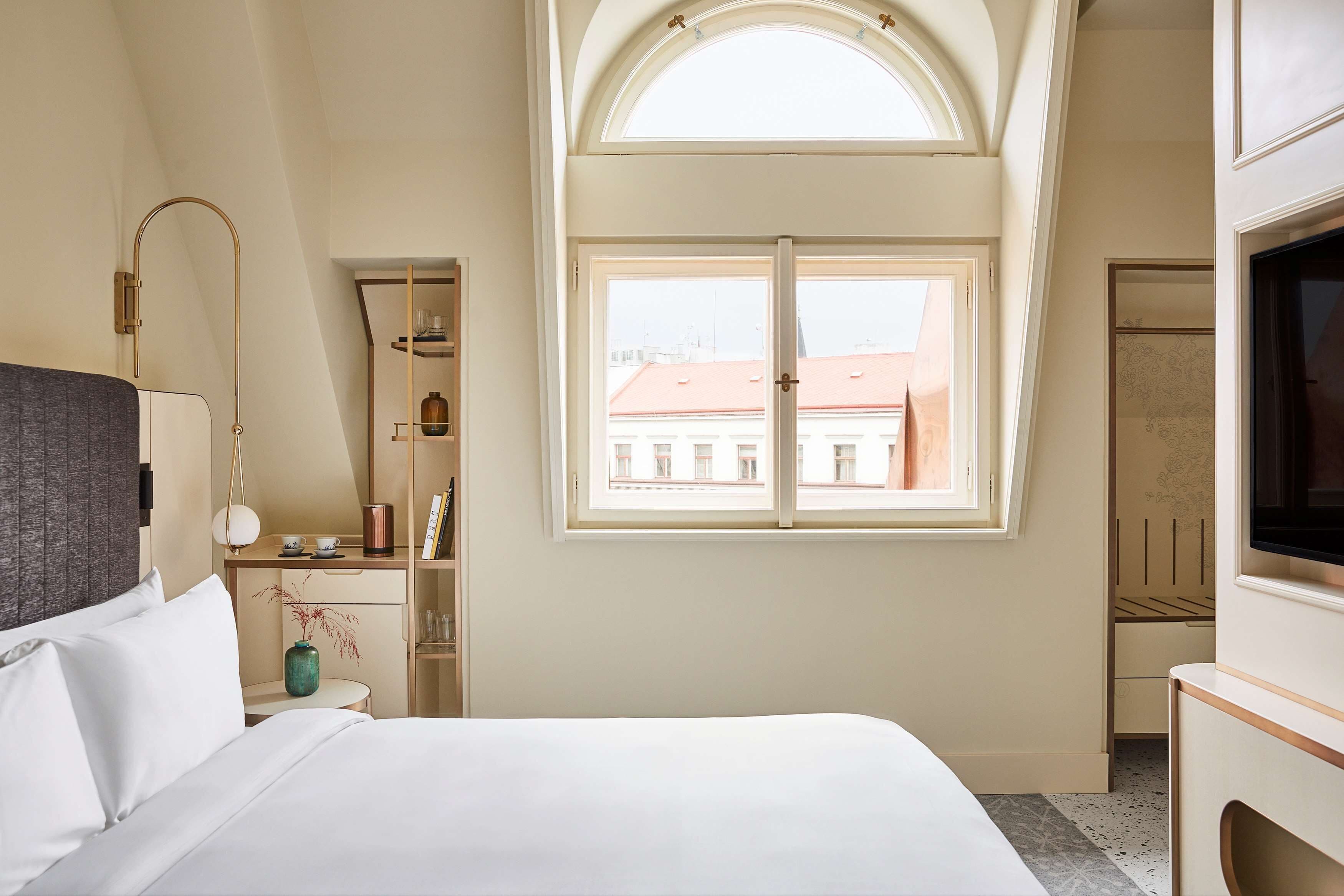 A hotel room with a bed, a desk, a chair, a TV, a window with a view of buildings, and a shelf with items.