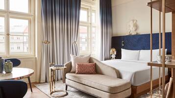 A hotel room with a bed, a sofa, a small table with a vase, and a view of buildings outside.
