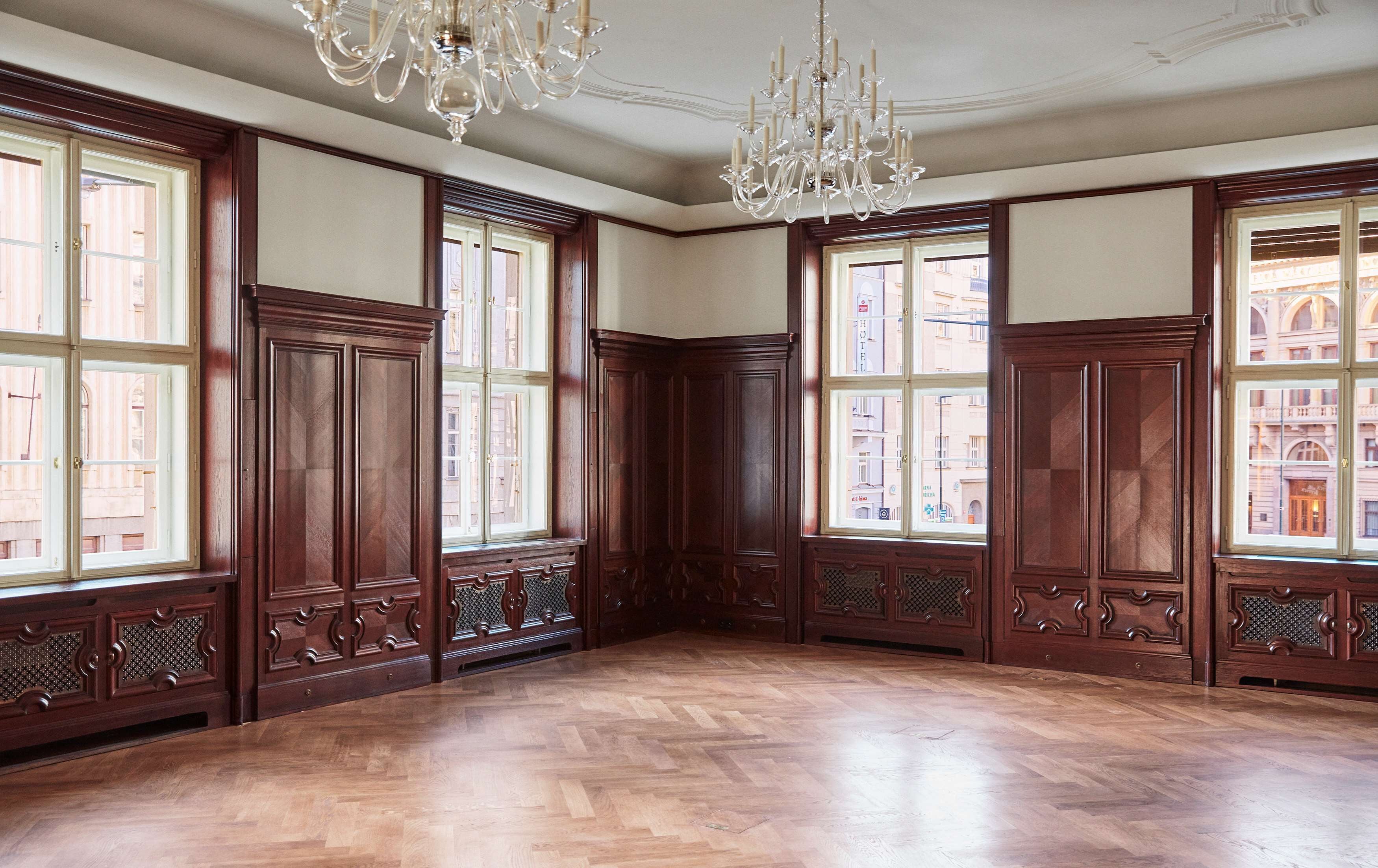 A spacious room with large wooden paneled walls, a herringbone wood floor, and a large window offering a view of buildings outside.