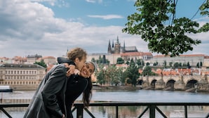 A couple embracing by a river with a historic bridge and buildings in the background. - ANDAZ PRAGUE, BY HYATT (Prague)