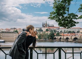A couple embracing by a river with a historic bridge and buildings in the background.