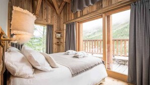 Une chambre dans une cabane en bois, avec un grand lit, un plaid blanc moelleux et une vue sur une terrasse en bois et les montagnes.