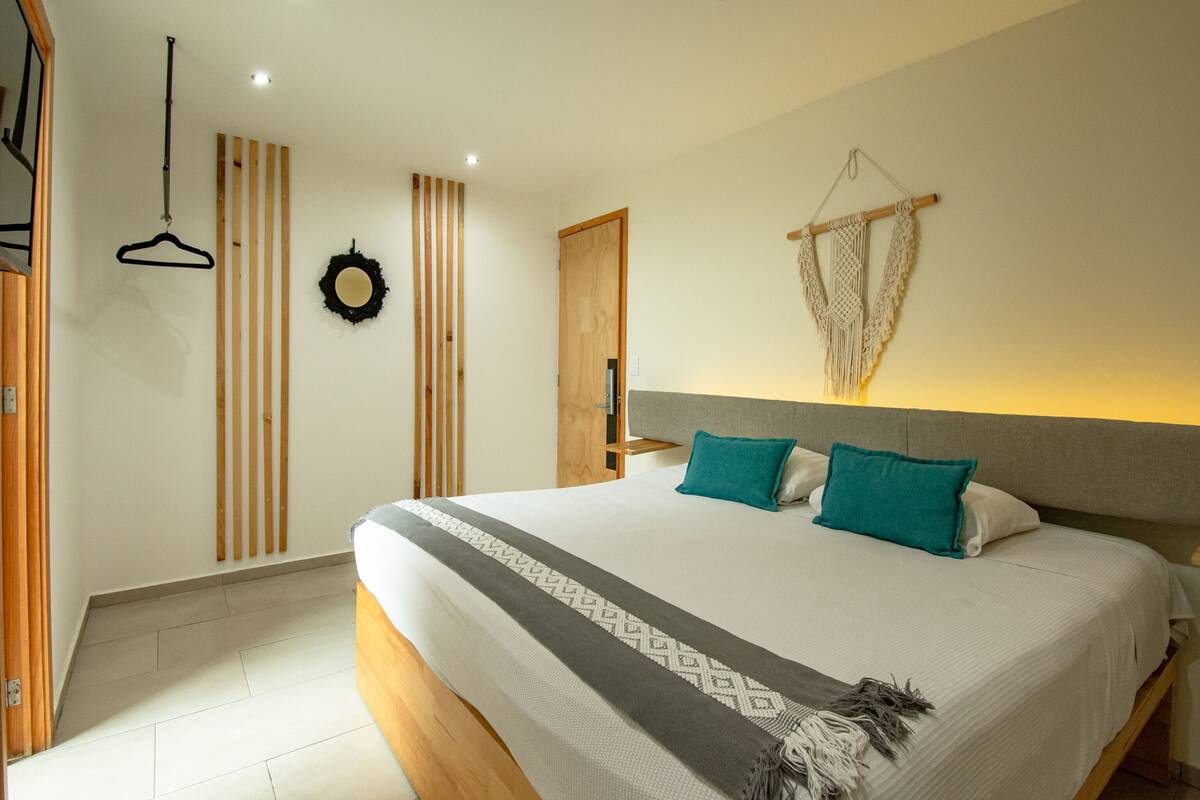 A bedroom with a large bed, wooden floor, a wooden door, a wall-mounted macrame, and a hanging black hanger.