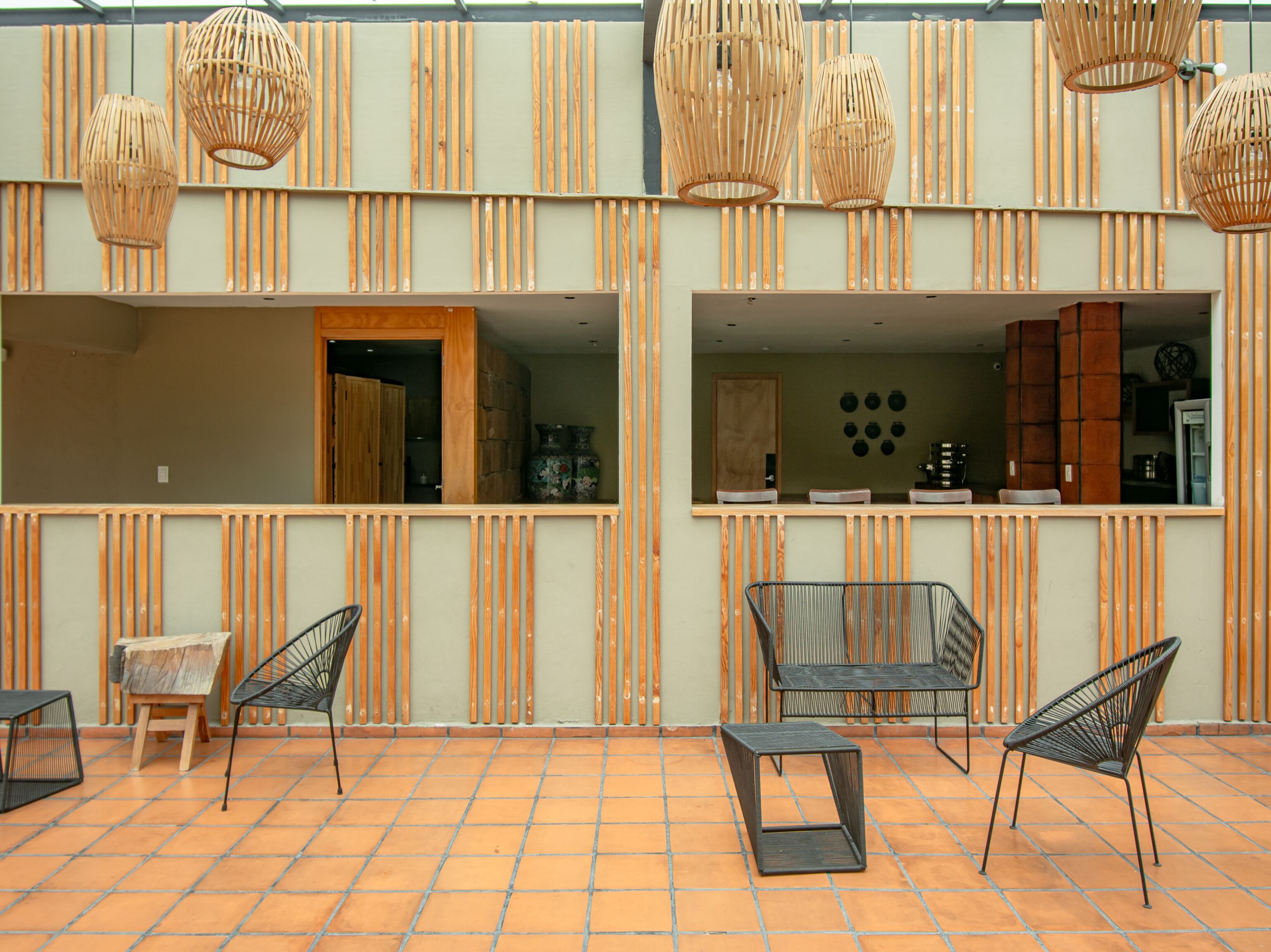 A modern outdoor seating area with wicker pendant lights, terracotta tiles, and a view of the interior through large windows.