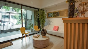 A reception area with a wooden reception desk, a round coffee table, two yellow chairs, a large window with a view of the street, and a potted plant. - Capital O Hotel Terraza 158 Mexico City (Mexico City)