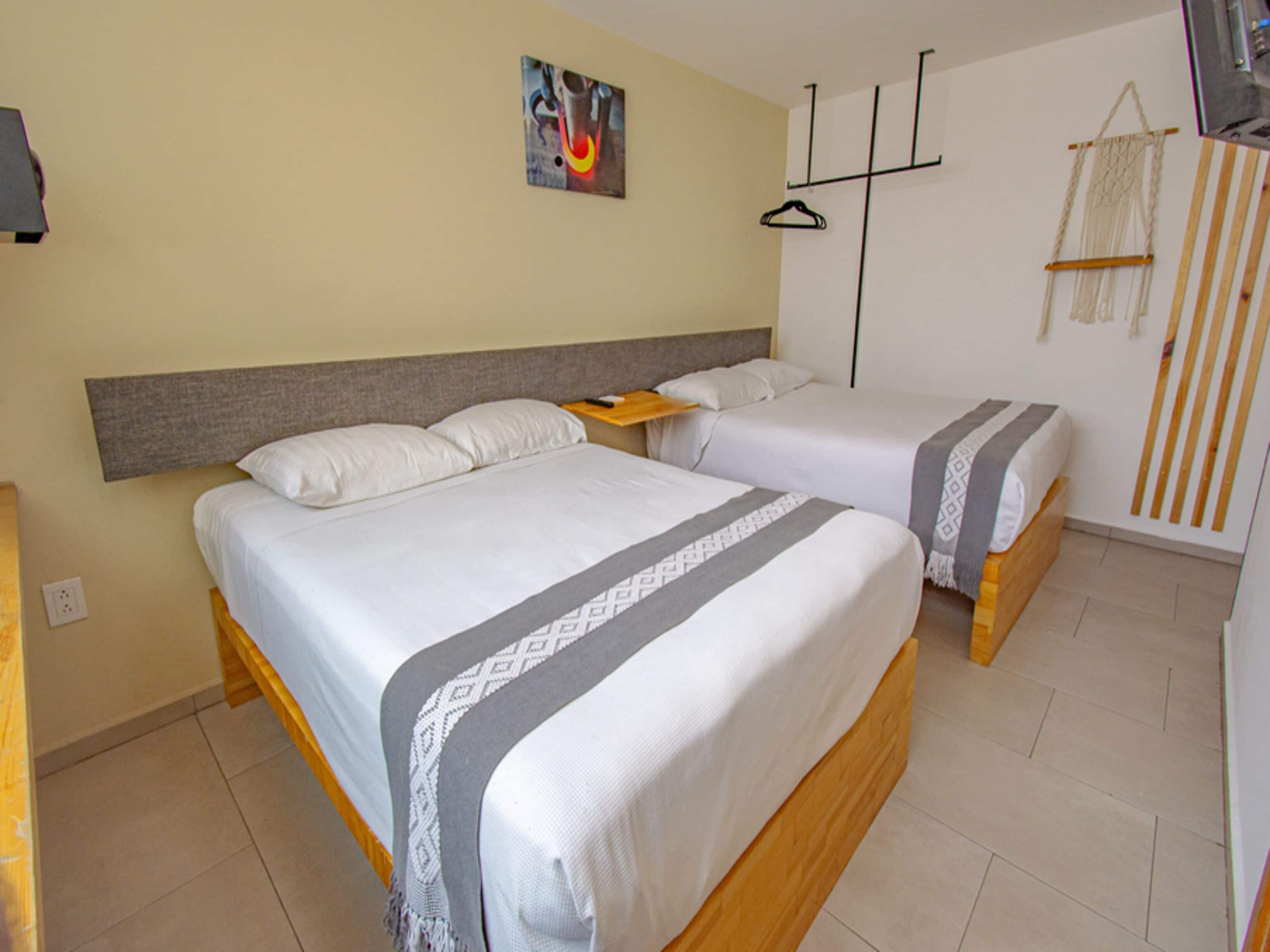 A hotel room with two single beds, a wooden headboard, a painting on the wall, a wooden side table, and a macrame wall hanging.