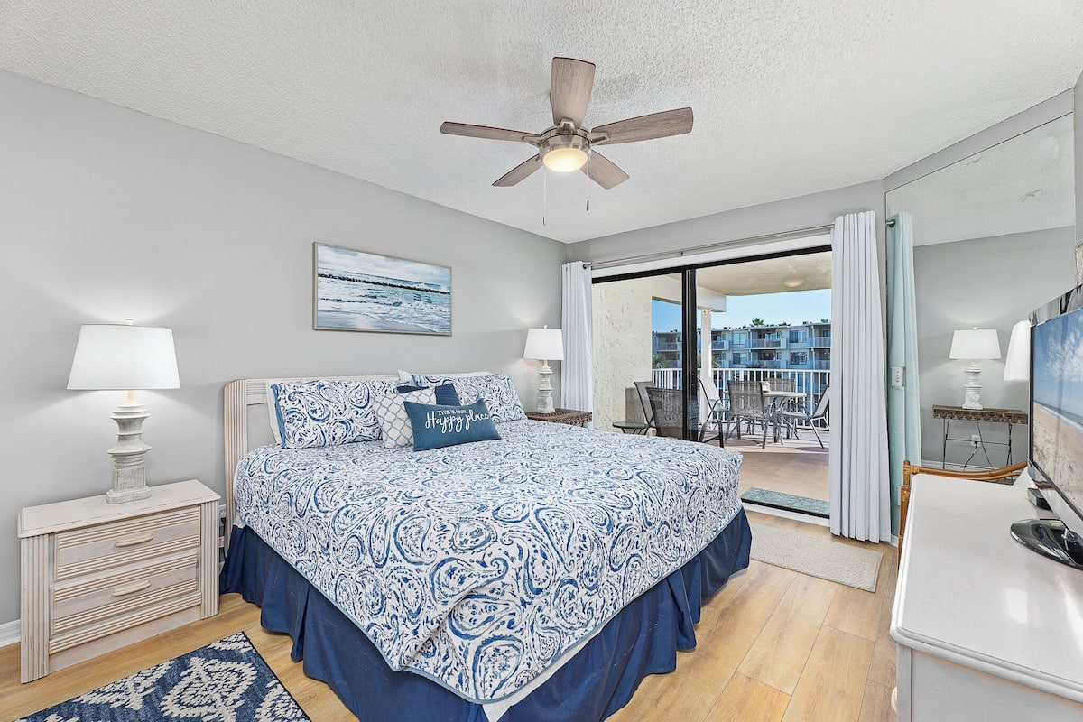 A bedroom with a bed, bedside lamps, a ceiling fan, a view of the balcony, and a painting on the wall.