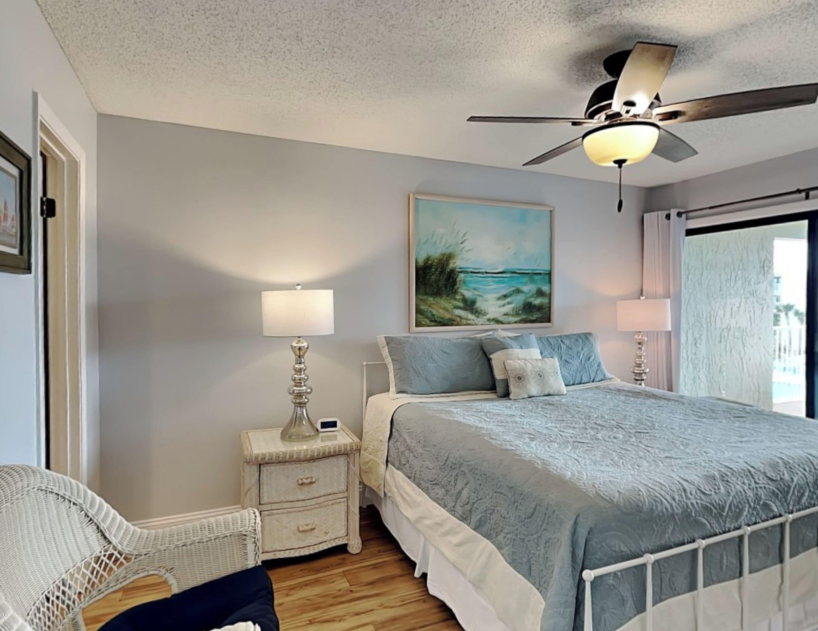 A bedroom with a bed, nightstands, a ceiling fan, and a painting of a beach scene.