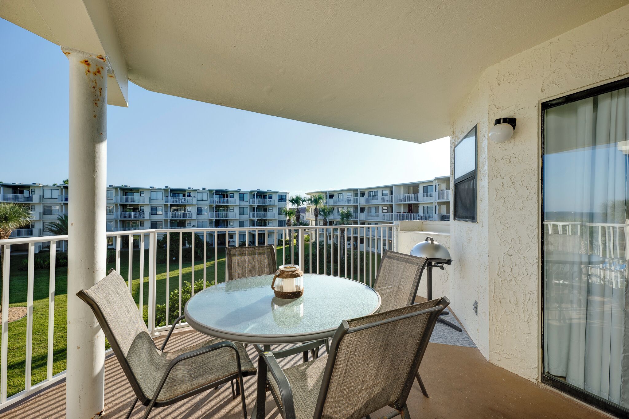 A balcony with a round glass table and four chairs, overlooking a residential area with multiple buildings.