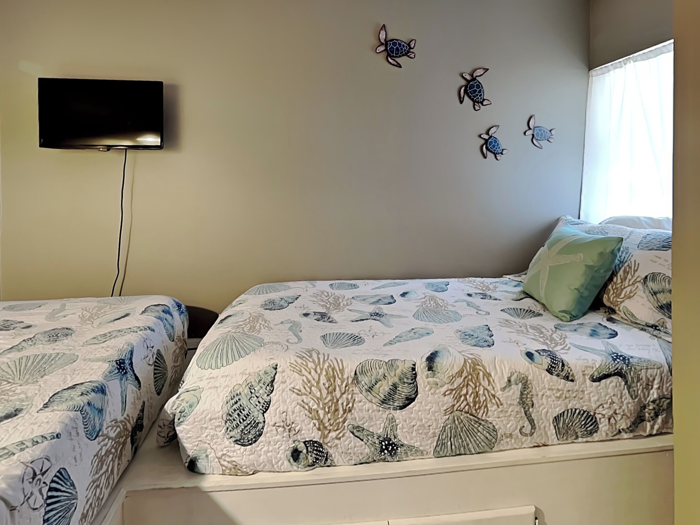 Ein Schlafzimmer mit zwei Betten, einem Flachbildfernseher an der Wand und einem Fenster mit durchsichtigen Vorhängen.