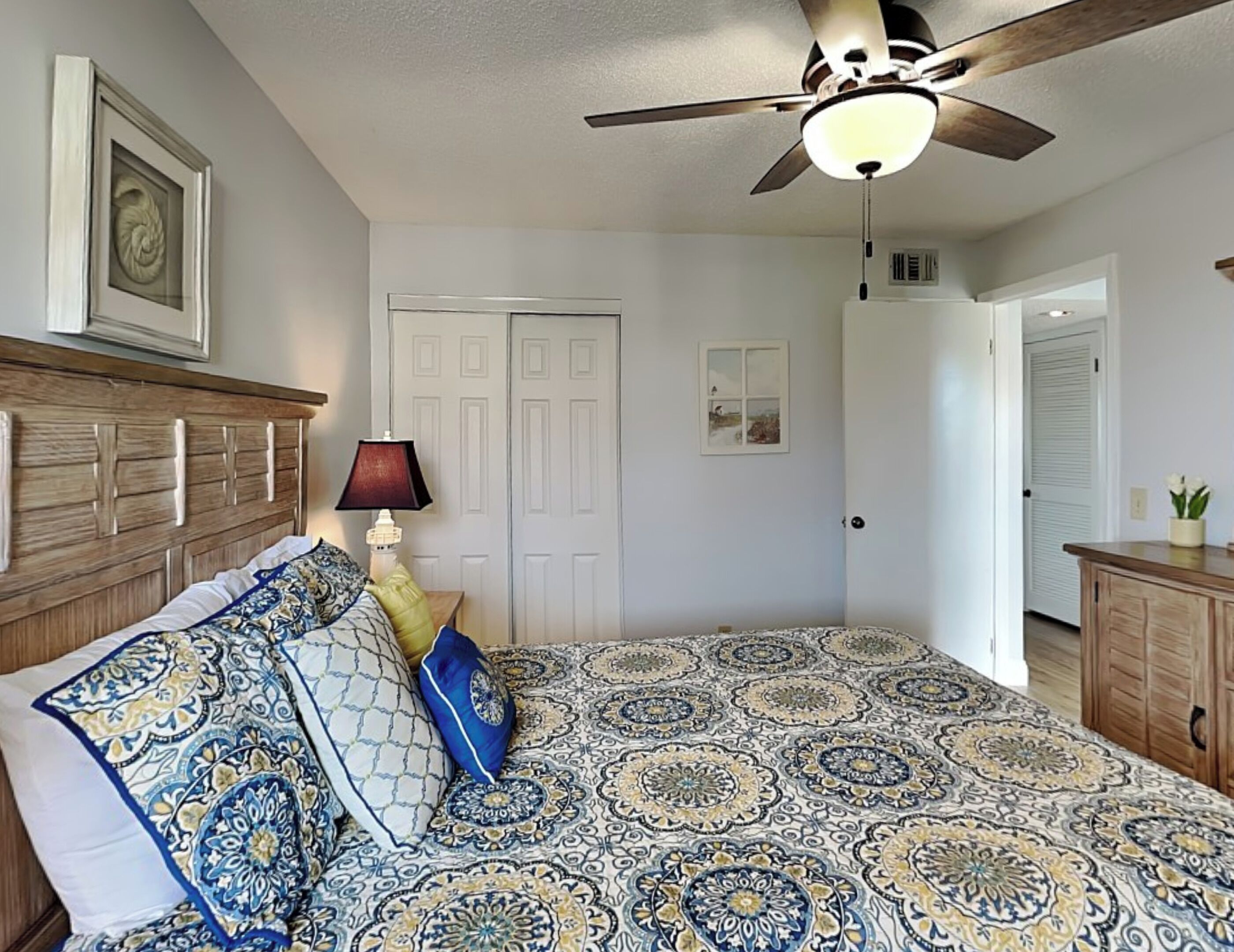 Une chambre à coucher avec une couvre-lit à motifs, une tête de lit en bois, un ventilateur de plafond et un tableau encadré au mur.
