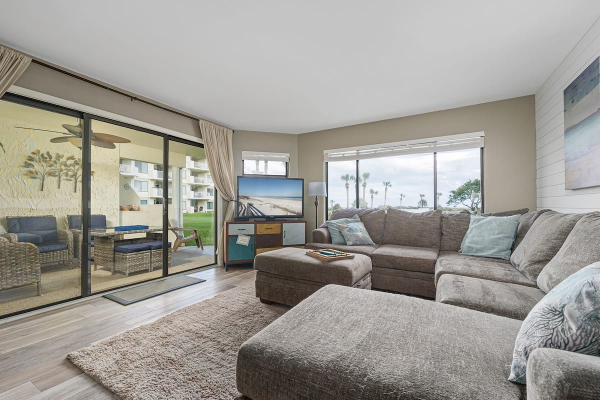 A living room with a large sectional sofa, a flat-screen TV, and a sliding glass door leading to a balcony.