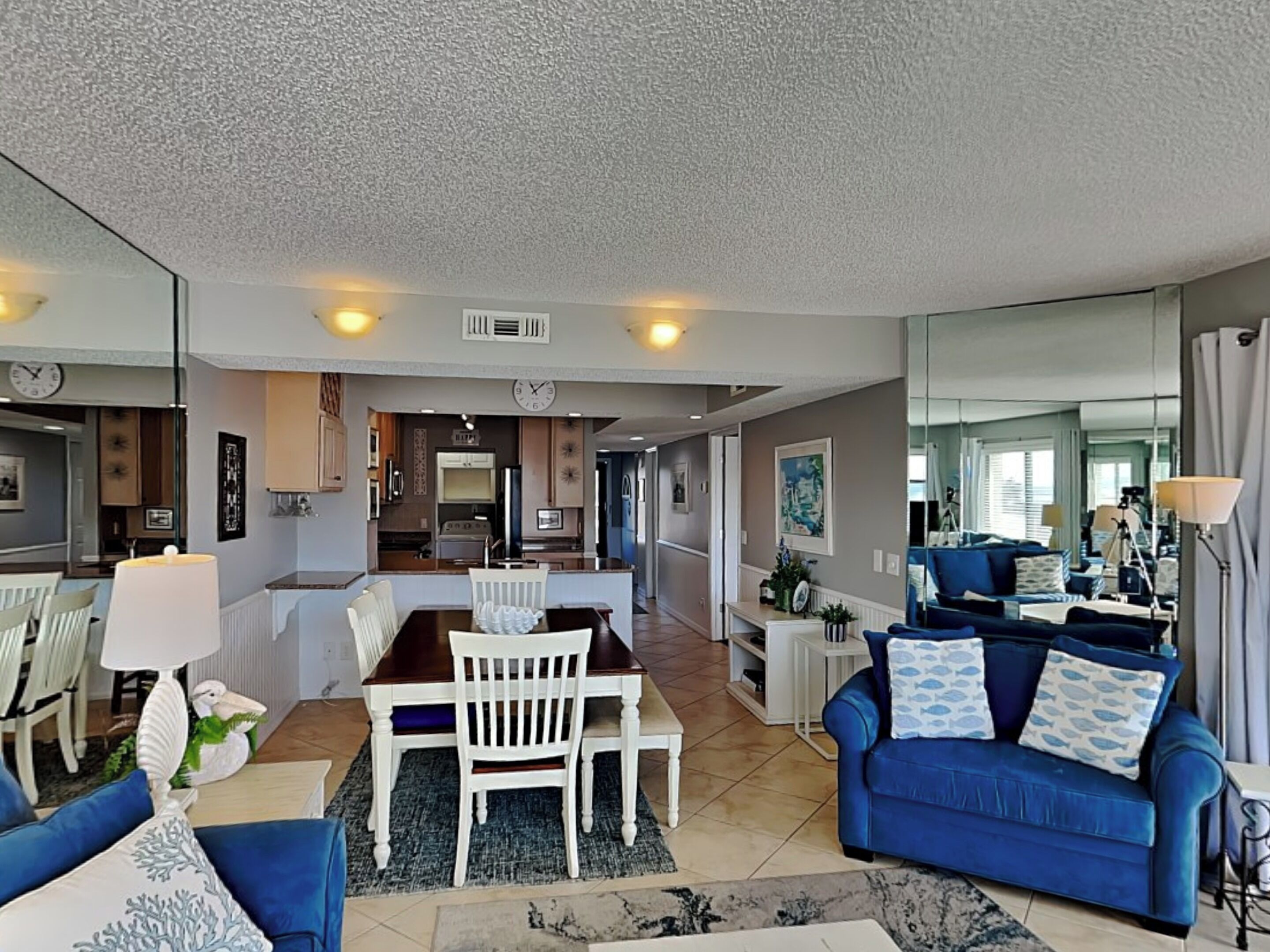 A modern living room with a blue sofa, a dining table with chairs, a kitchen area, and a large mirror reflecting the space.