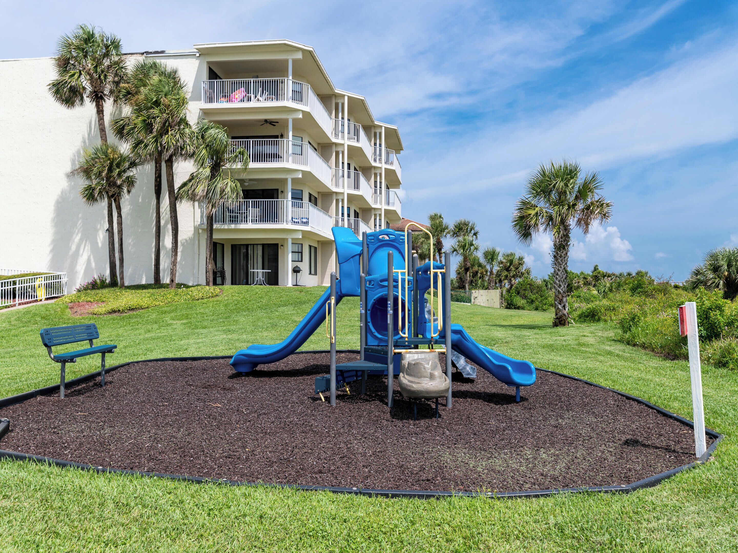 Ein Spielplatz mit einer blauen Rutsche und einer Kletterkonstruktion, umgeben von einem gepflegten Rasen und Palmen, mit einem mehrstöckigen Gebäude im Hintergrund.