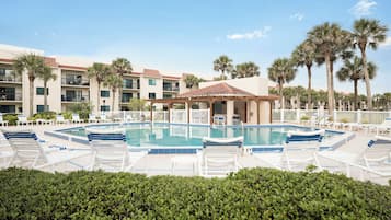 A pool area with lounge chairs and a gazebo, surrounded by palm trees and a multi-story building with balconies.