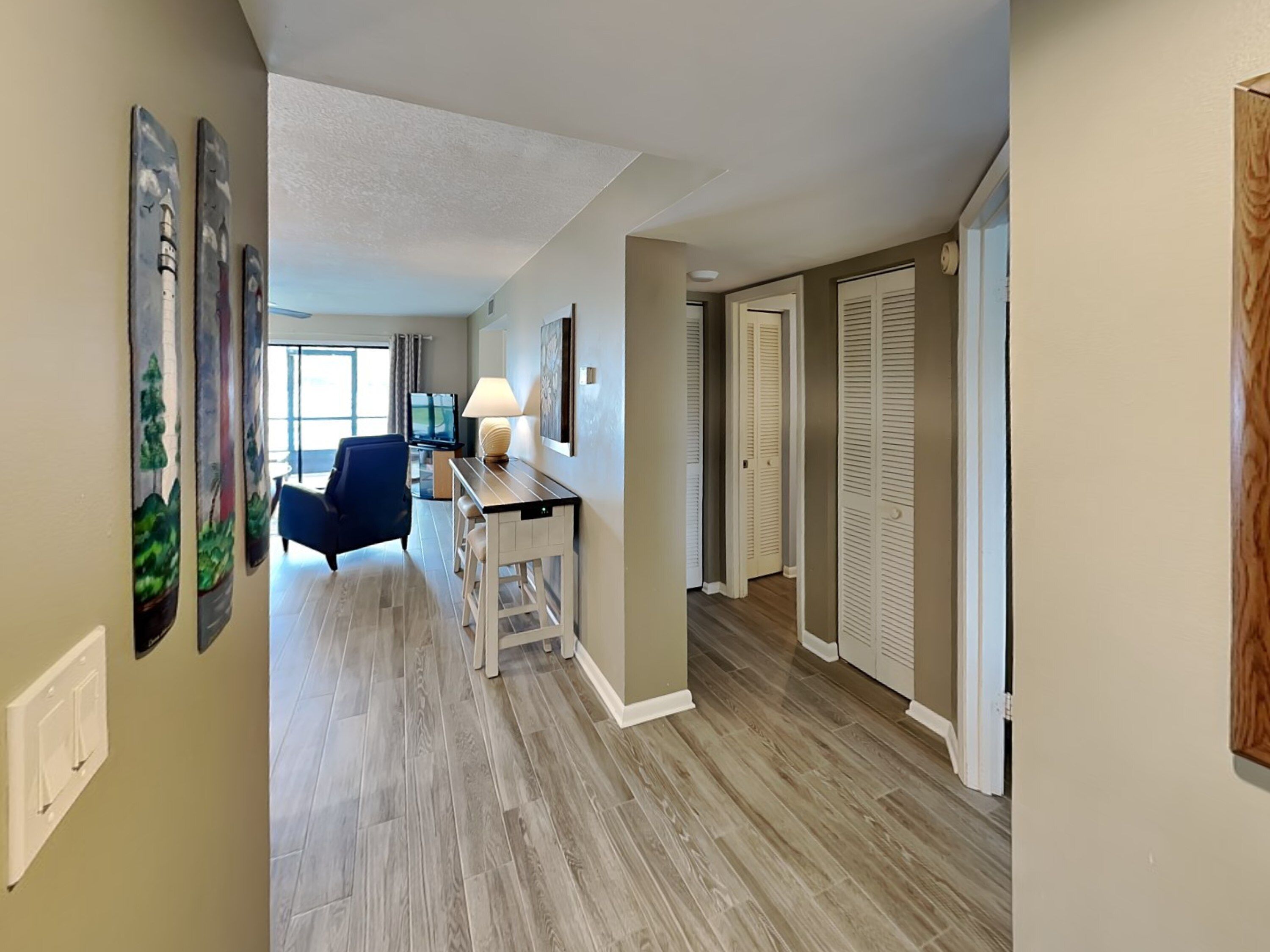 Un couloir d’hôtel avec un plancher en bois, un bureau avec une lampe, une chaise bleue et un téléviseur.