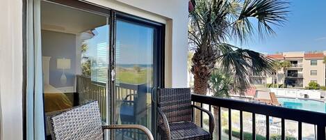A balcony with wicker chairs, a glass door leading to a room, and a view of a pool and palm trees.