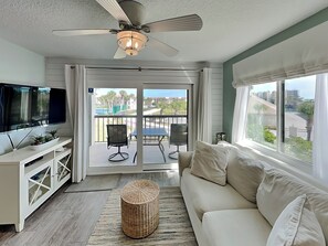 A living room with a TV, sofa, and a balcony with chairs. - Ocean Village Club by Distinctive Beach Rentals (St. Augustine)