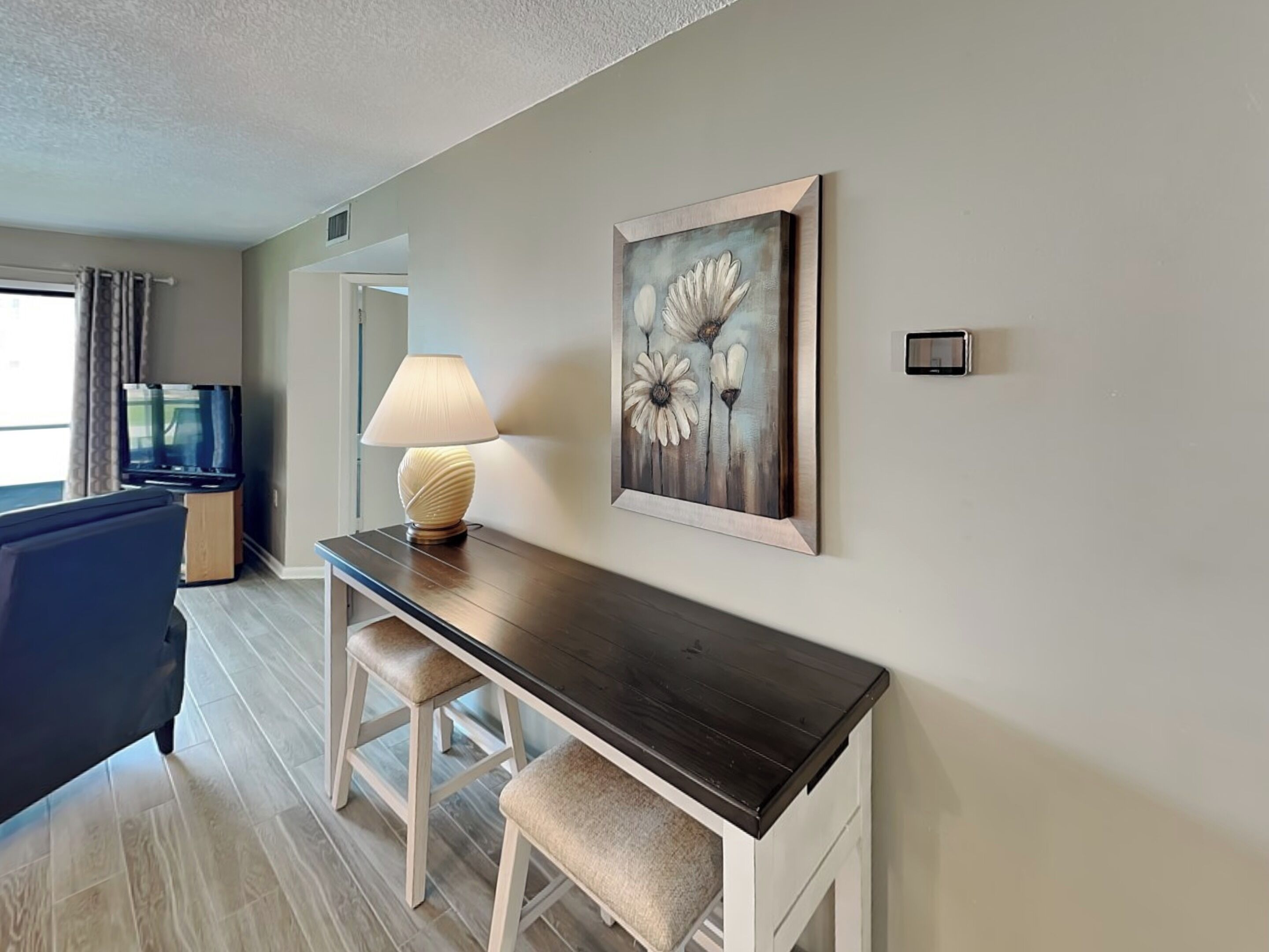 A modern living room with a dark wooden coffee table, two white cushioned stools, a beige sofa, and a television on a wooden stand. A painting of flowers hangs on the wall.