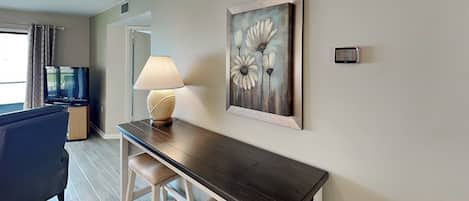 Una sala de estar moderna con una mesa de centro de madera oscura, dos taburetes blancos acolchados, un sofá beige y un televisor sobre un mueble de madera. En la pared, un cuadro con flores.