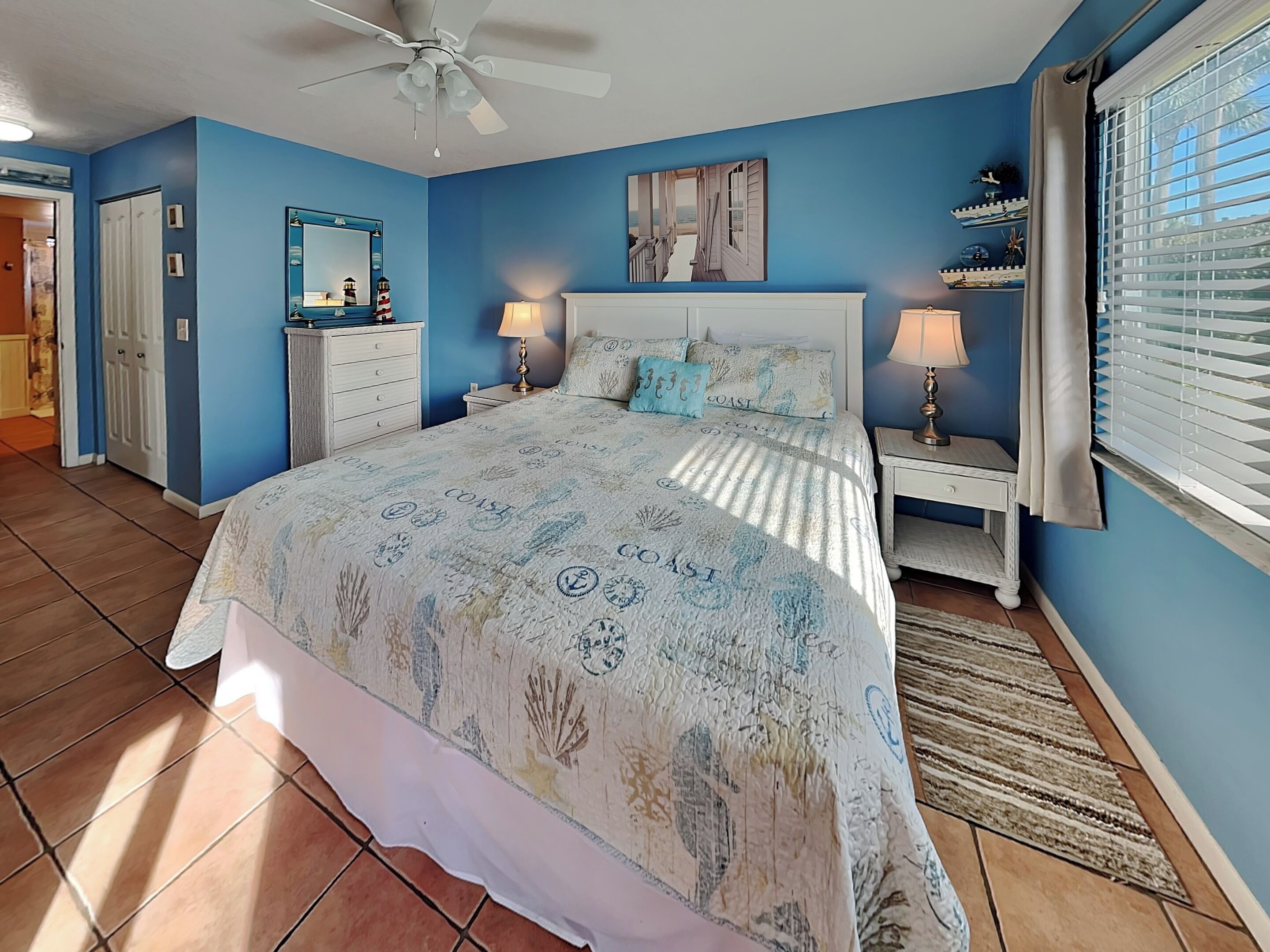 A bedroom with a bed, two bedside tables, a ceiling fan, a mirror, and a window with blinds.