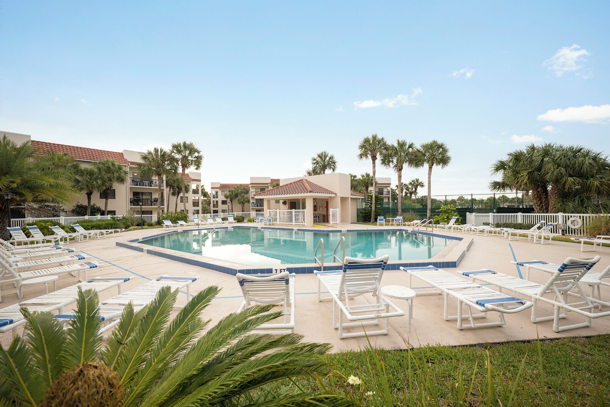 A pool area with lounge chairs, palm trees, and a building with a red-tiled roof.