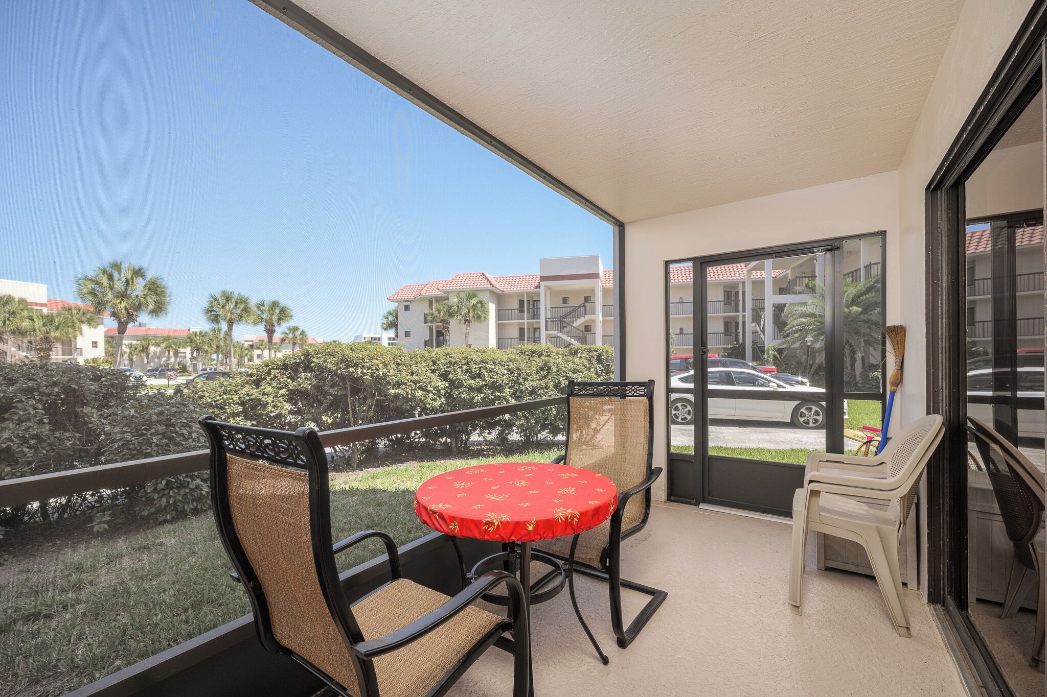 A balcony with a table and chairs, a view of a street with buildings and palm trees, and a door leading outside.