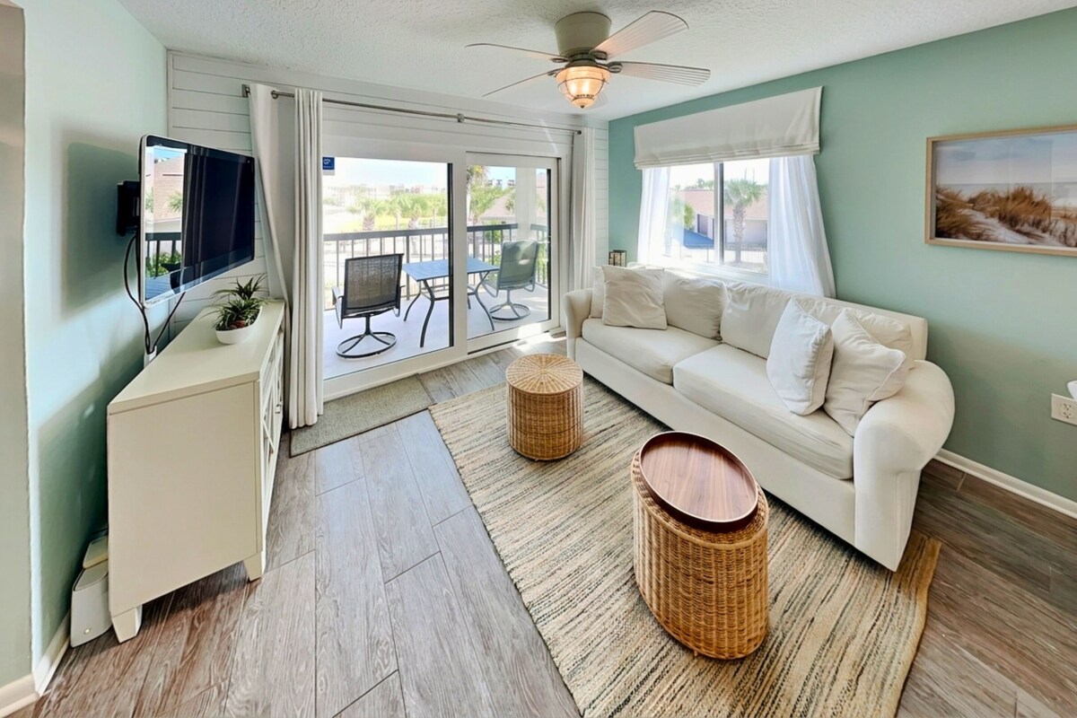 A living room with a white sofa, a TV, a ceiling fan, and a view through a sliding glass door to an outdoor patio.