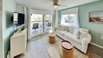 A living room with a white sofa, a TV, a ceiling fan, and a view through a sliding glass door to an outdoor patio.