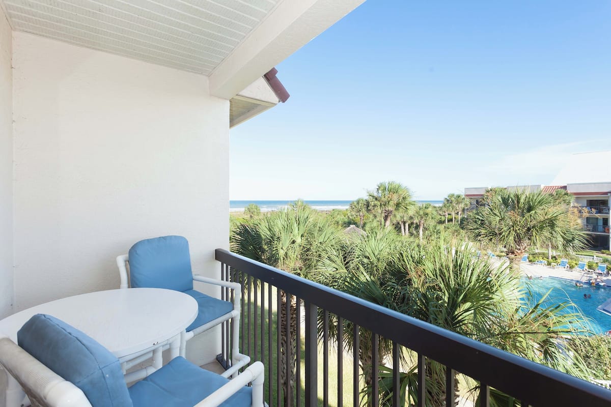 Ein Balkon mit einem weißen Tisch und blauen Stühlen, der auf einen Pool und Palmen blickt.