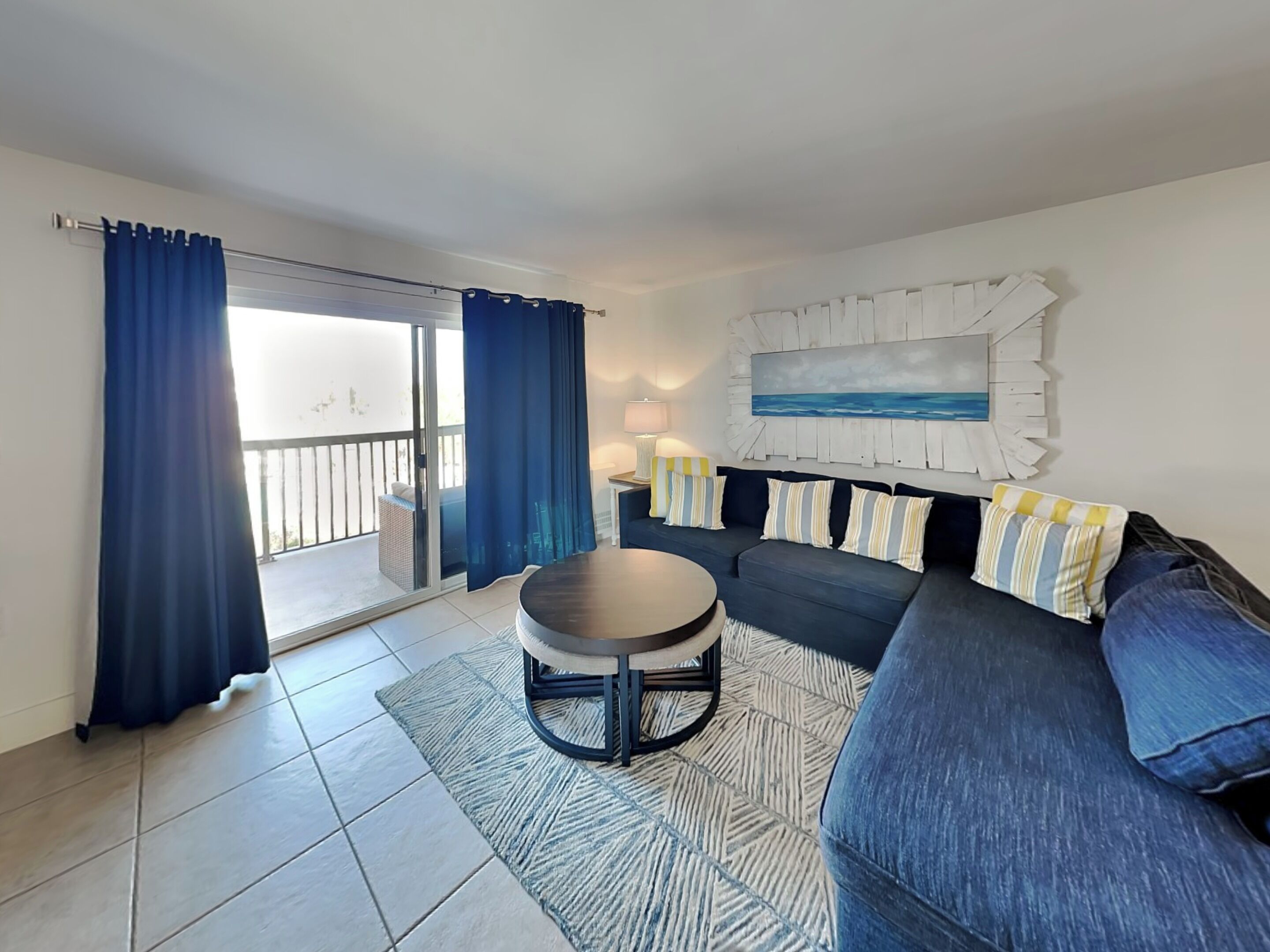 A living room with a dark blue sofa, a round coffee table, and a patterned rug. There is a sliding glass door leading to a balcony.