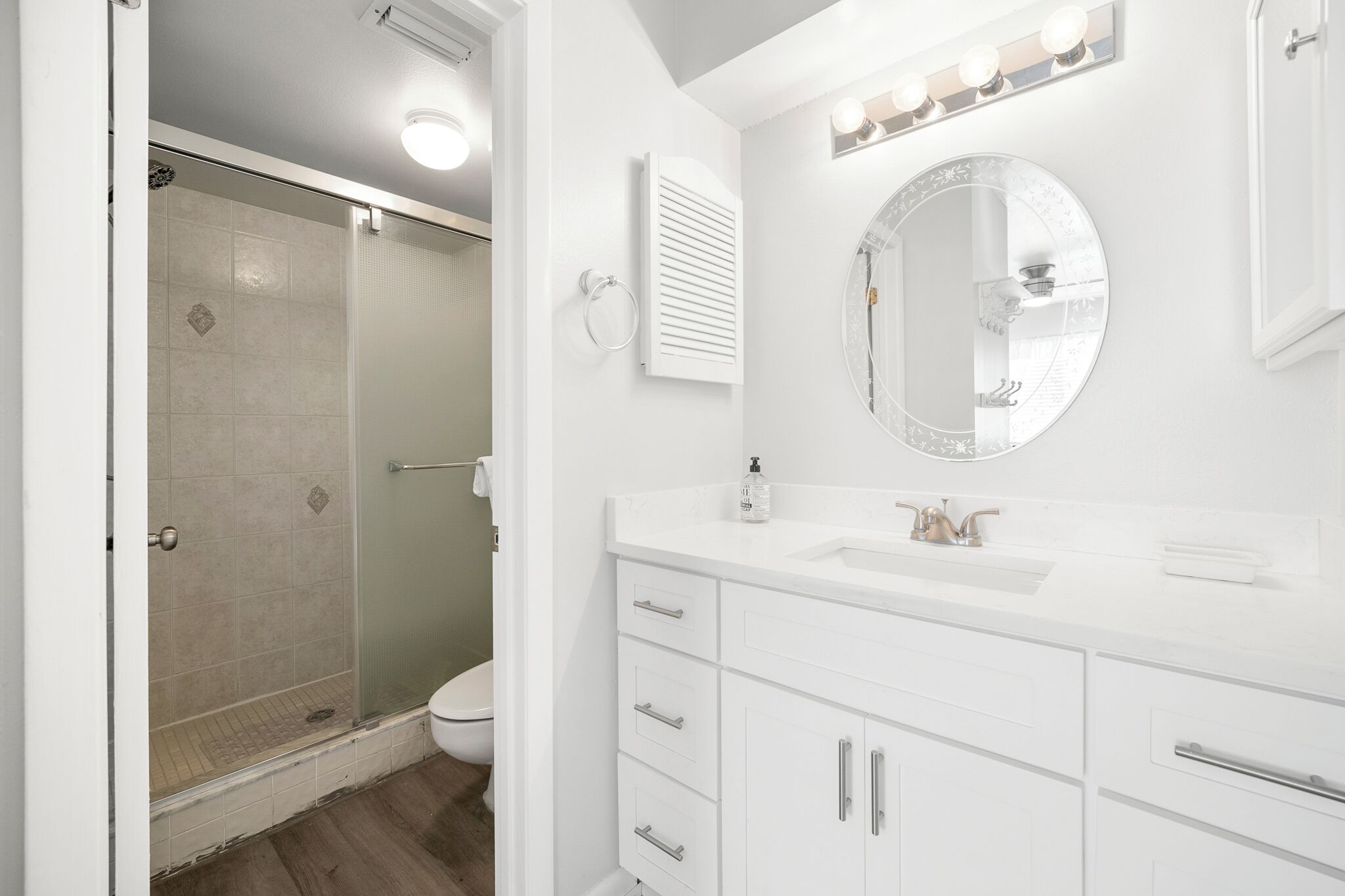 Une salle de bain avec une douche à parois de verre, un miroir rond et des meubles de rangement blancs.