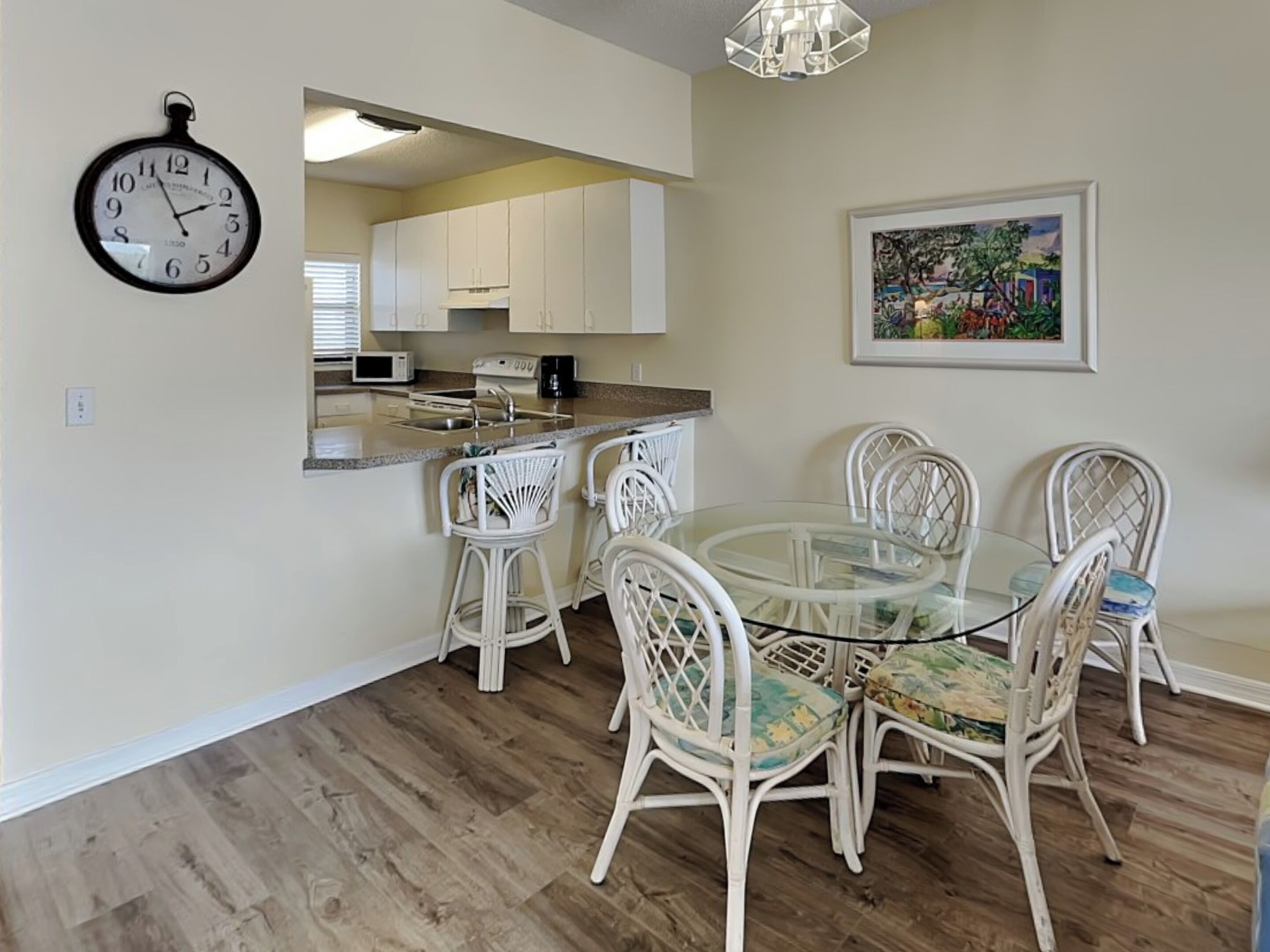 A dining area with a glass table and white wicker chairs, a kitchen with white cabinets, and a wall clock.
