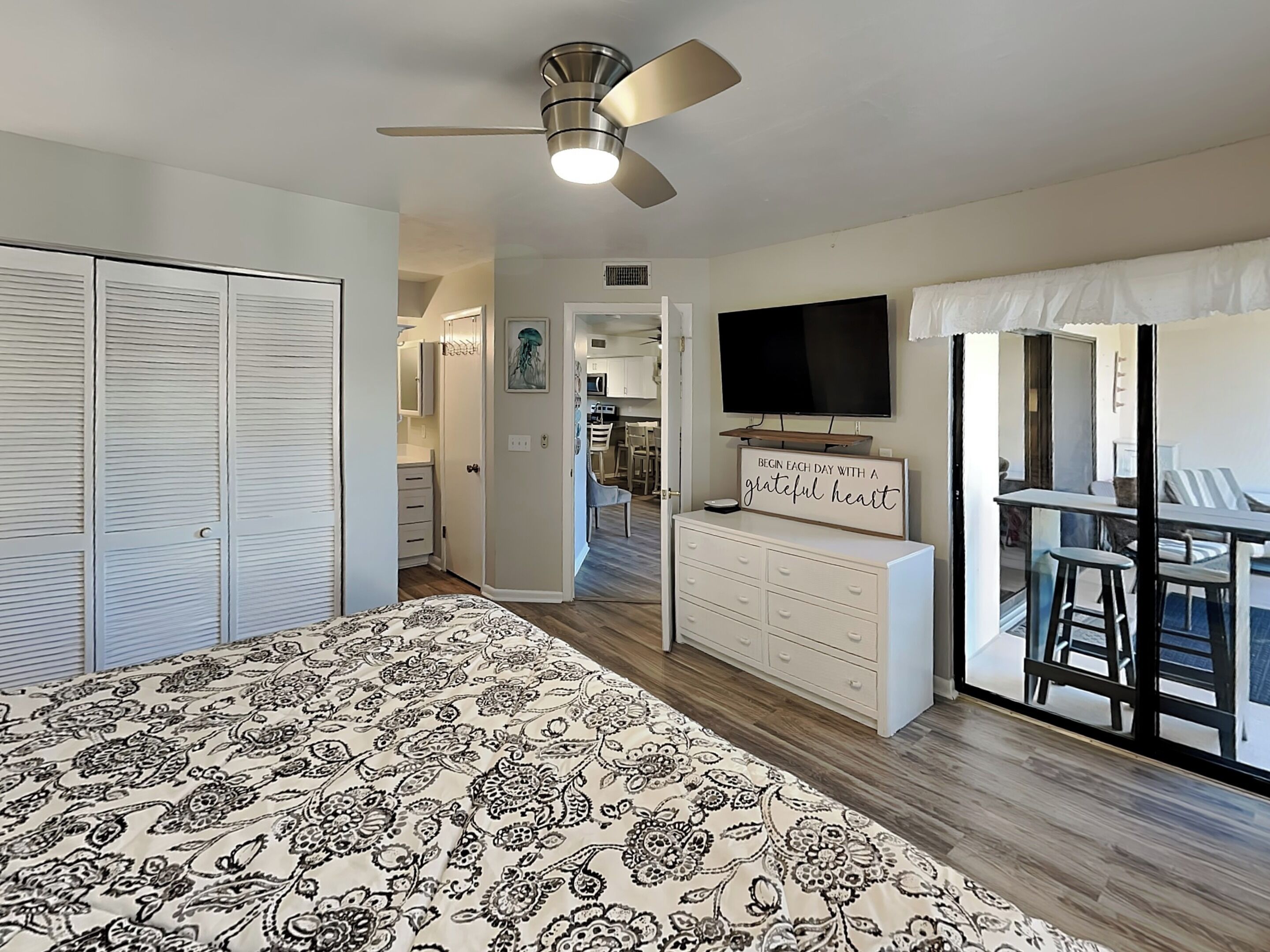 A bedroom with a bed, a ceiling fan, a dresser, a TV, and a balcony with chairs.