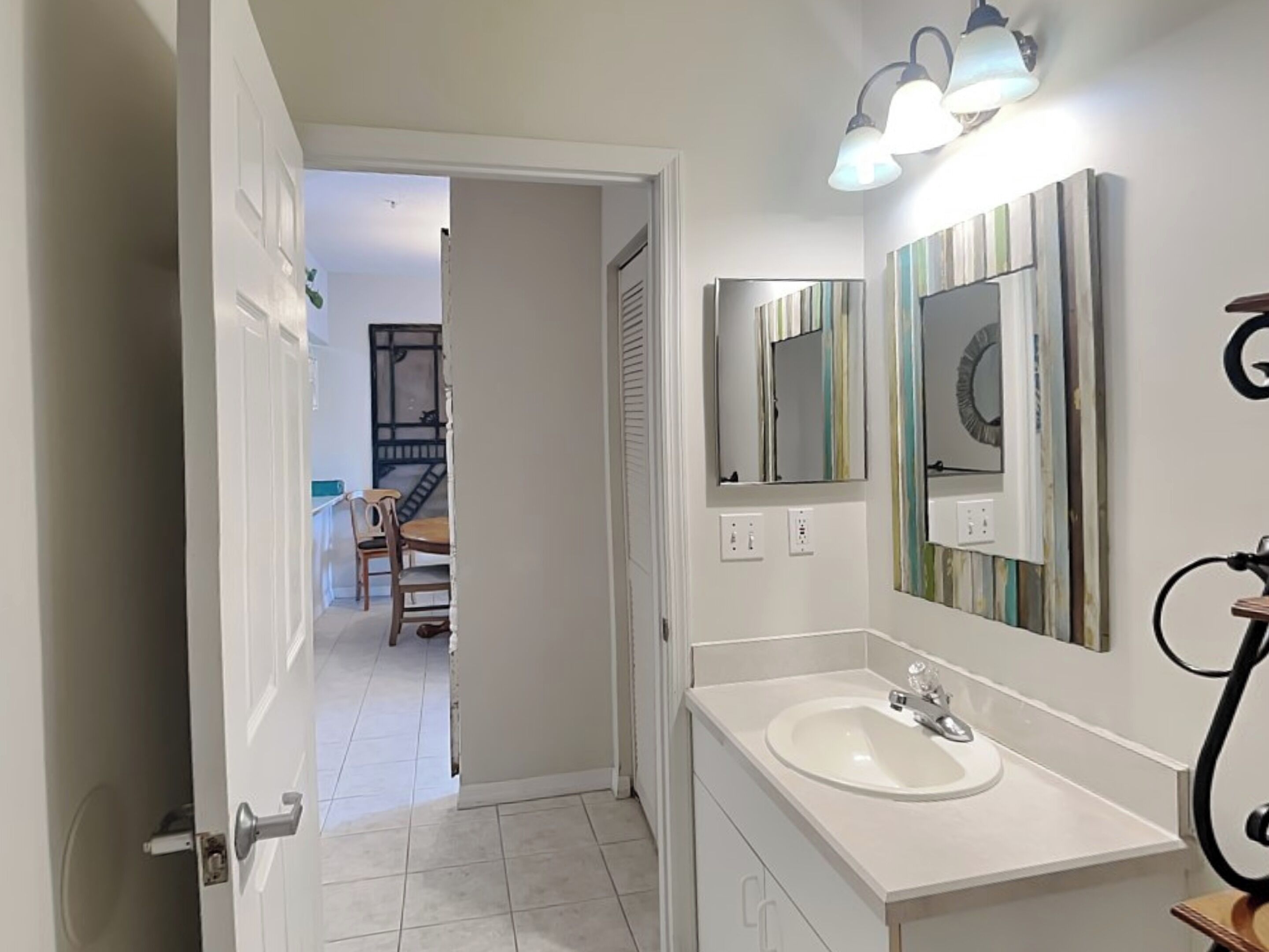 Une salle de bain avec un lavabo blanc, un miroir et une douche. Une porte donne sur un couloir où se trouve un escalier.