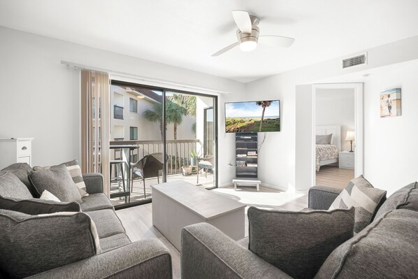 A modern living room with a sofa set, a flat-screen TV, and a balcony with a view of a building and palm trees. - Ocean Village Club by Distinctive Beach Rentals (St. Augustine)