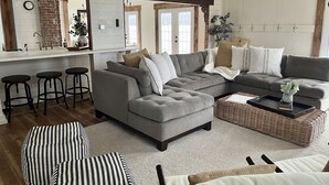 A modern living room with a grey sectional sofa, a coffee table, and striped ottomans. There is a kitchen area with a white countertop and stools. - Birchwood Cottage, 200ft of Lake Beach-Fully flat lot -Private Driveway (Branchport)