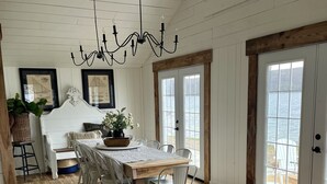 A dining area with a wooden table and chairs, a chandelier, and a view of the outdoors through large windows. - Birchwood Cottage, 200ft of Lake Beach-Fully flat lot -Private Driveway (Branchport)