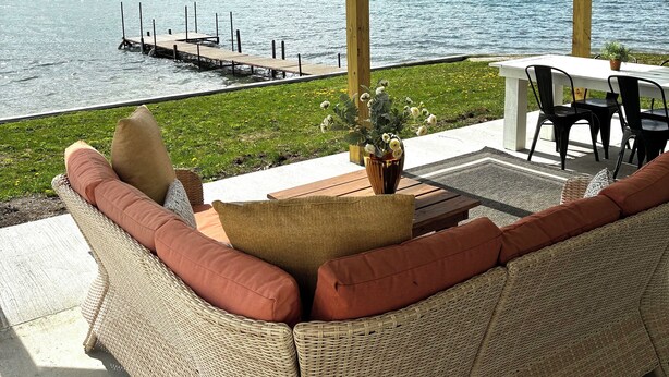 A covered outdoor seating area with wicker furniture, a table, and chairs, overlooking a lake with a dock. - Birchwood Cottage, 200ft of Lake Beach-Fully flat lot -Private Driveway (Branchport)