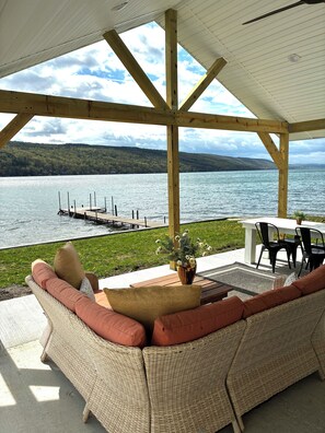 A covered outdoor seating area with wicker furniture, a table, and chairs, overlooking a lake with a dock. - Birchwood Cottage, 200ft of Lake Beach-Fully flat lot -Private Driveway (Branchport)
