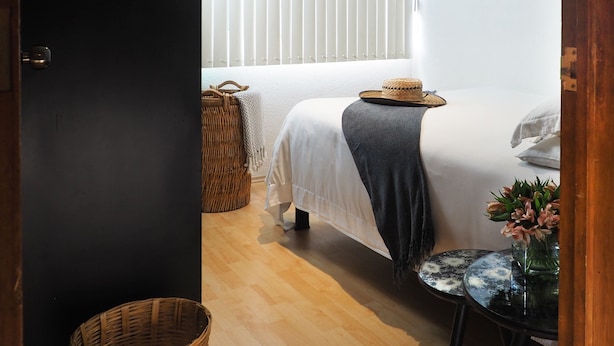 Ein Schlafzimmer mit einem Bett, einem Nachttisch mit Blumenvase, einem Korbwägelchen und einem Fenster mit Jalousien.