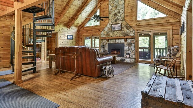 A log cabin interior with a stone fireplace, a spiral staircase, and a leather sofa.