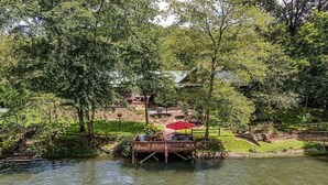 A wooden deck over a river with a table and chairs, surrounded by trees and a grassy area.