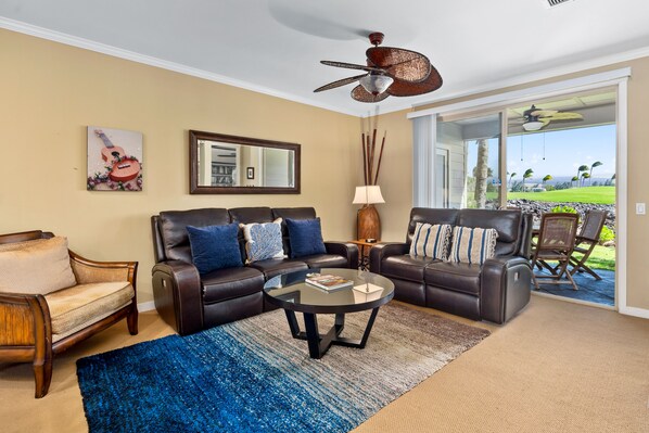A living room with a brown leather sofa, a glass coffee table, and a large window offering a view of a green landscape. - Hilton Waikoloa Pool Pass for stays thru 2026. ML Beach Club Pass. Golf VillasMa (Mauna Lani Resort)