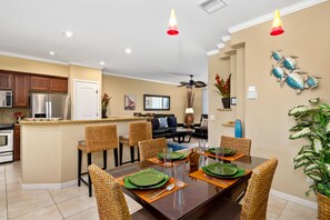 A modern kitchen with stainless steel appliances, a dining area with wicker chairs and a dark wooden table, and a living room with a sofa and coffee table. - Hilton Waikoloa Pool Pass for stays thru 2026. ML Beach Club Pass. Golf VillasMa (Mauna Lani Resort)